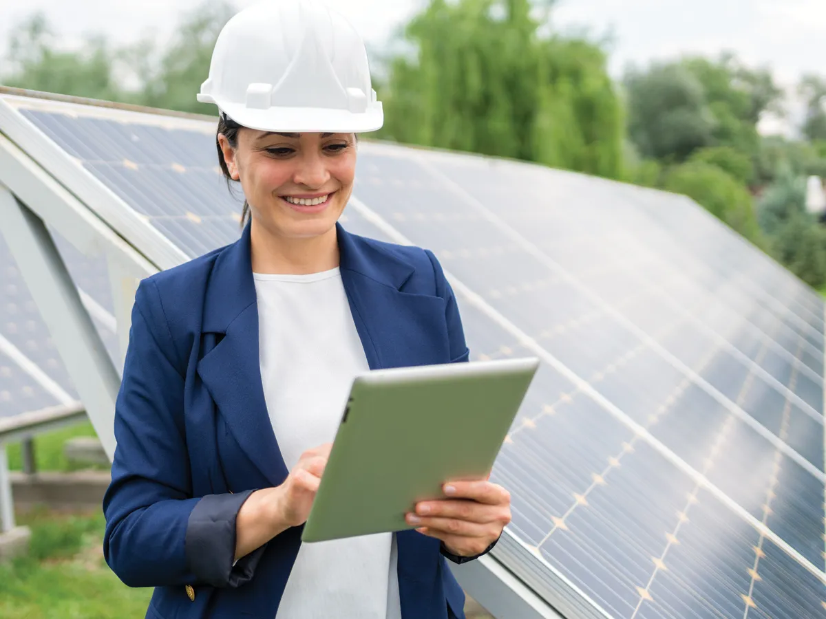 Professional woman wearing a hard hat and looking at a tablet. She's standing in front of ground-mount solar panels.