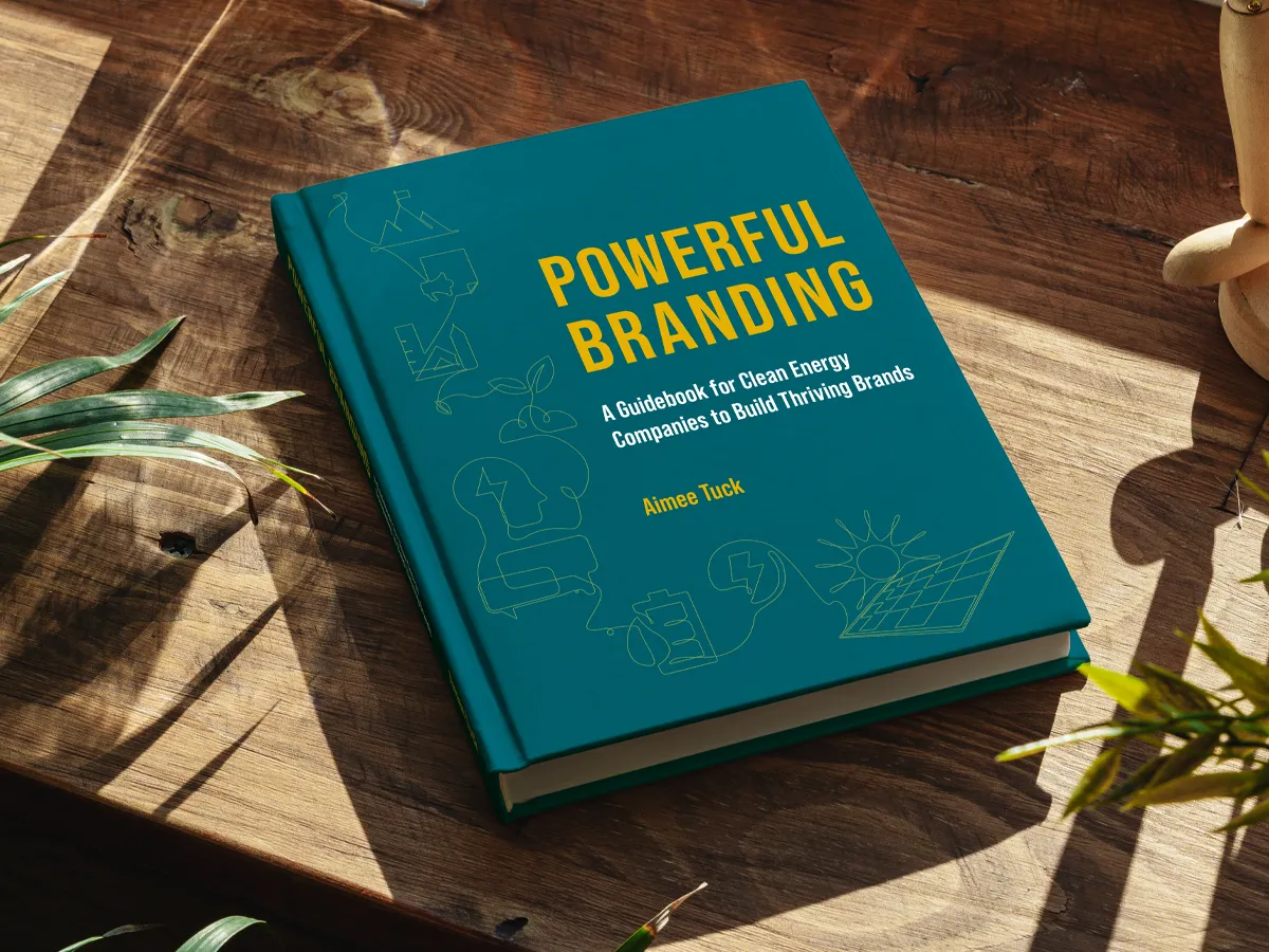 A book, titled Powerful Branding, A Guidebook for Clean Energy Companies to Build Thriving Brands by Aimee Tuck sits on a wood desk in the sunlight. Plants can be seen to either side of the book. 